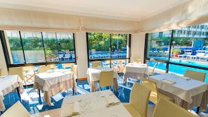 Restaurante com mesas preparadas para refeições, grandes janelas com vista para uma piscina e vegetação.