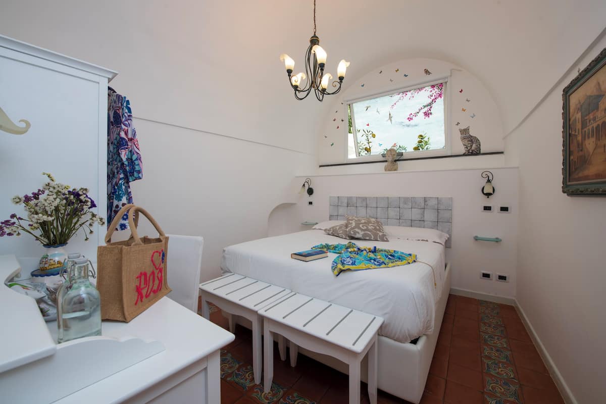 A bedroom with a bed, a bench, a desk, a chandelier, a window with a cat decoration, and a painting on the wall.