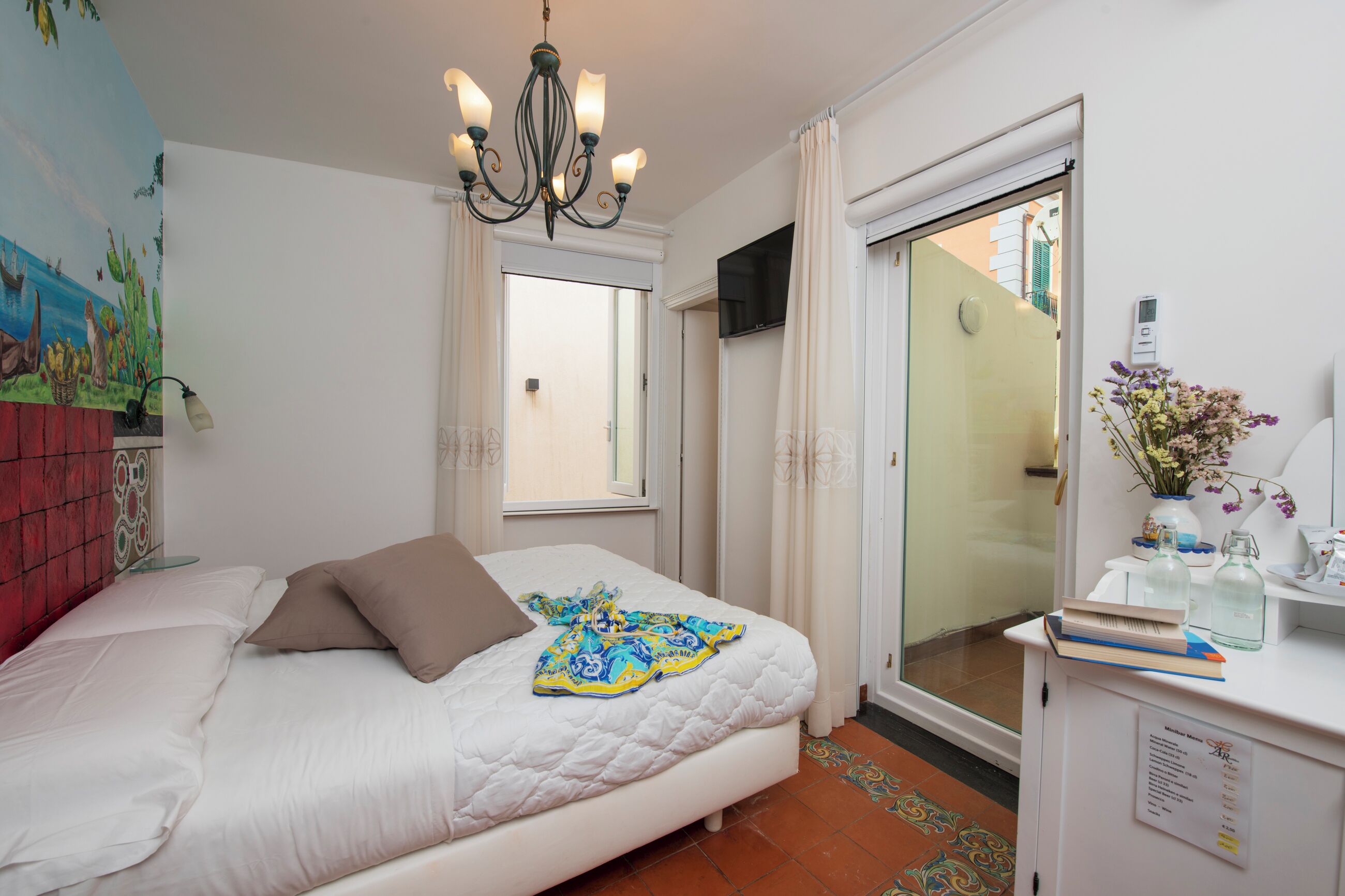 A bedroom with a bed, a chandelier, a TV, a window, and a desk with a vase of flowers.