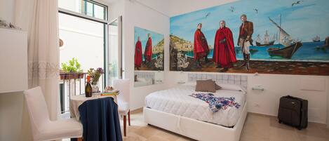 Un dormitorio con cama, una silla, una mesa con una botella y un cuadro en la pared.