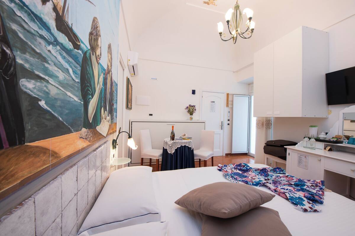 A hotel room with a large painting of a seascape, a bed with pillows, a desk with a chair, and a television on the wall.