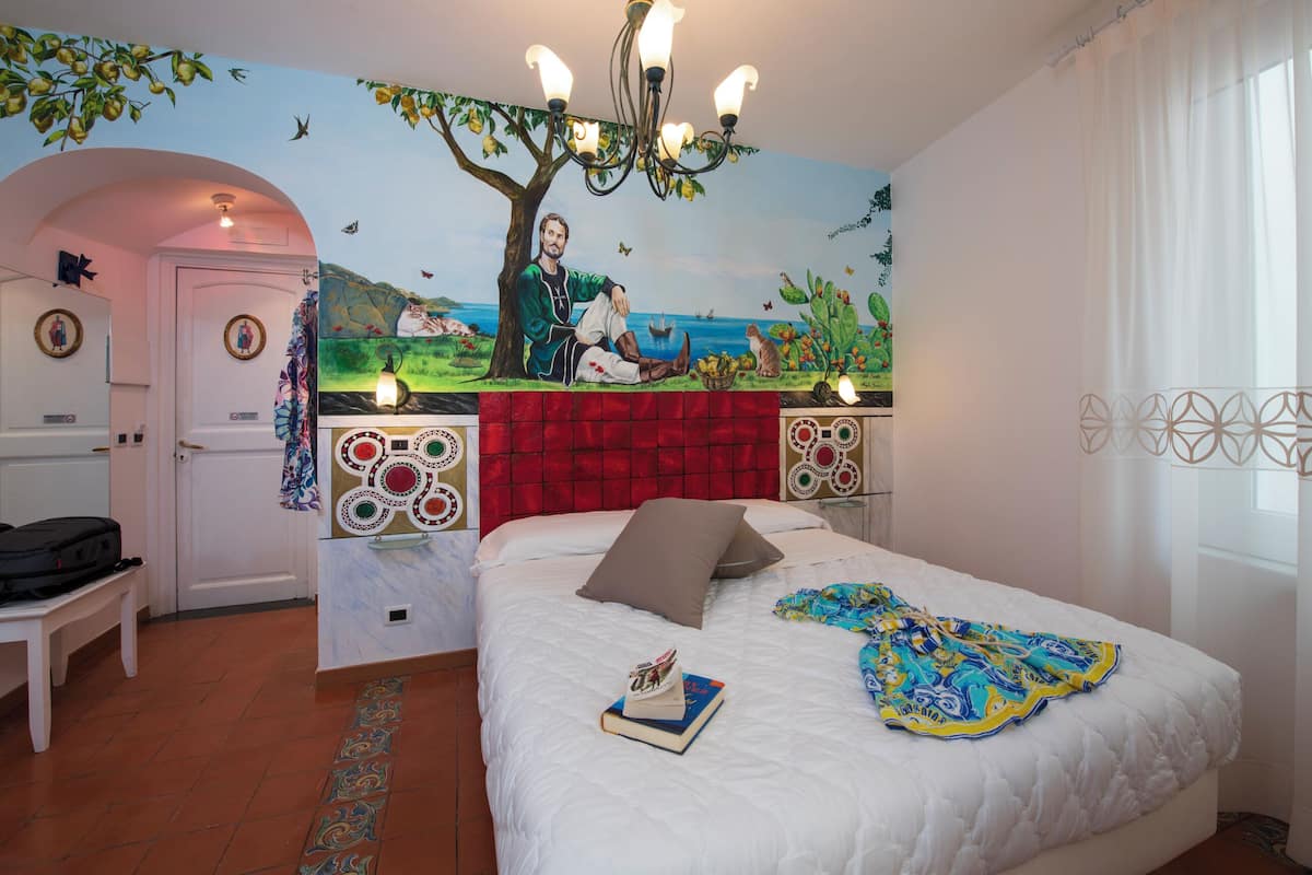 A bedroom with a mural of a man by a tree, a bed with a blue and yellow blanket, and a red headboard.