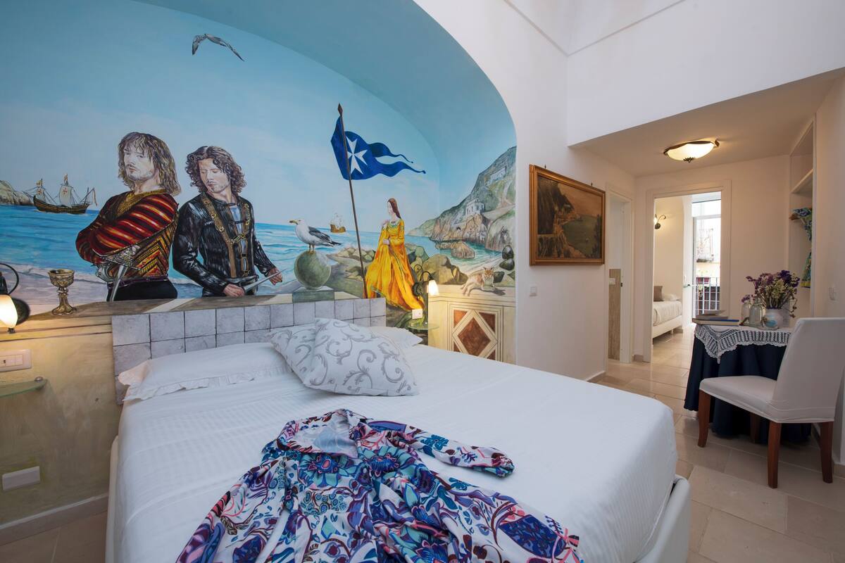A bedroom with a mural of a coastal scene, a bed with a patterned blanket, a chair, and a table with a vase.