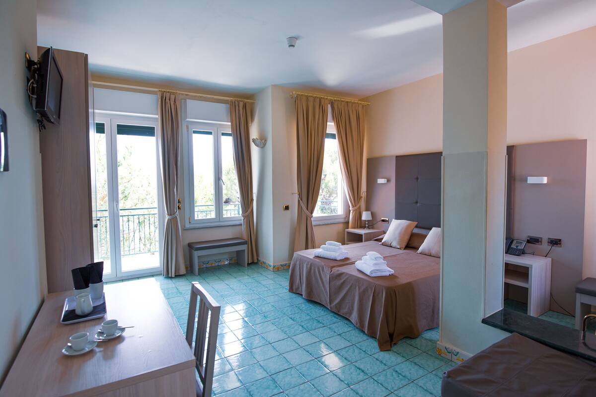 A hotel room with a bed, bedside table, and a desk with a chair. There is a window with curtains, a door, and a small table with a lamp.