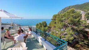 Terraza con mobiliario exterior que da al mar, barandilla y vistas a una ladera con árboles y un edificio.