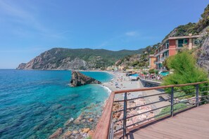 Vista costera con una terraza de madera que da a una playa, agua azul cristalina y edificios con balcones.