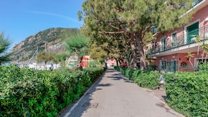 Un sendero bordeado de setos verdes y un gran pino, que conduce a un edificio con balcones y un autobús rojo al fondo.