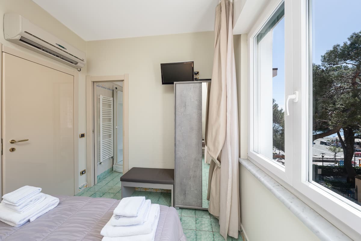 A hotel room with a bed, a bench, a television, and a window with a view of trees and buildings.