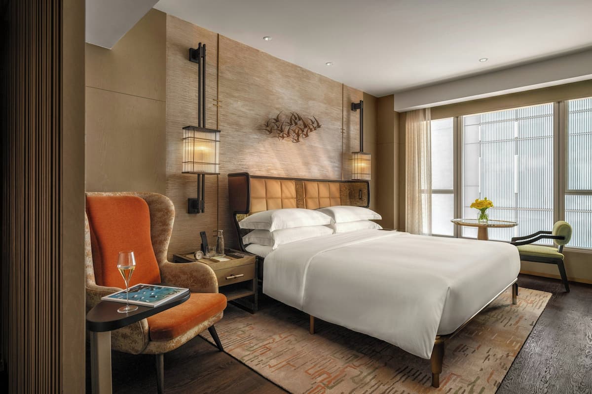 Een hotelkamer met een groot bed, een stoel, een tafeltje met een glas wijn, een nachtkastje en een lamp aan de muur.
