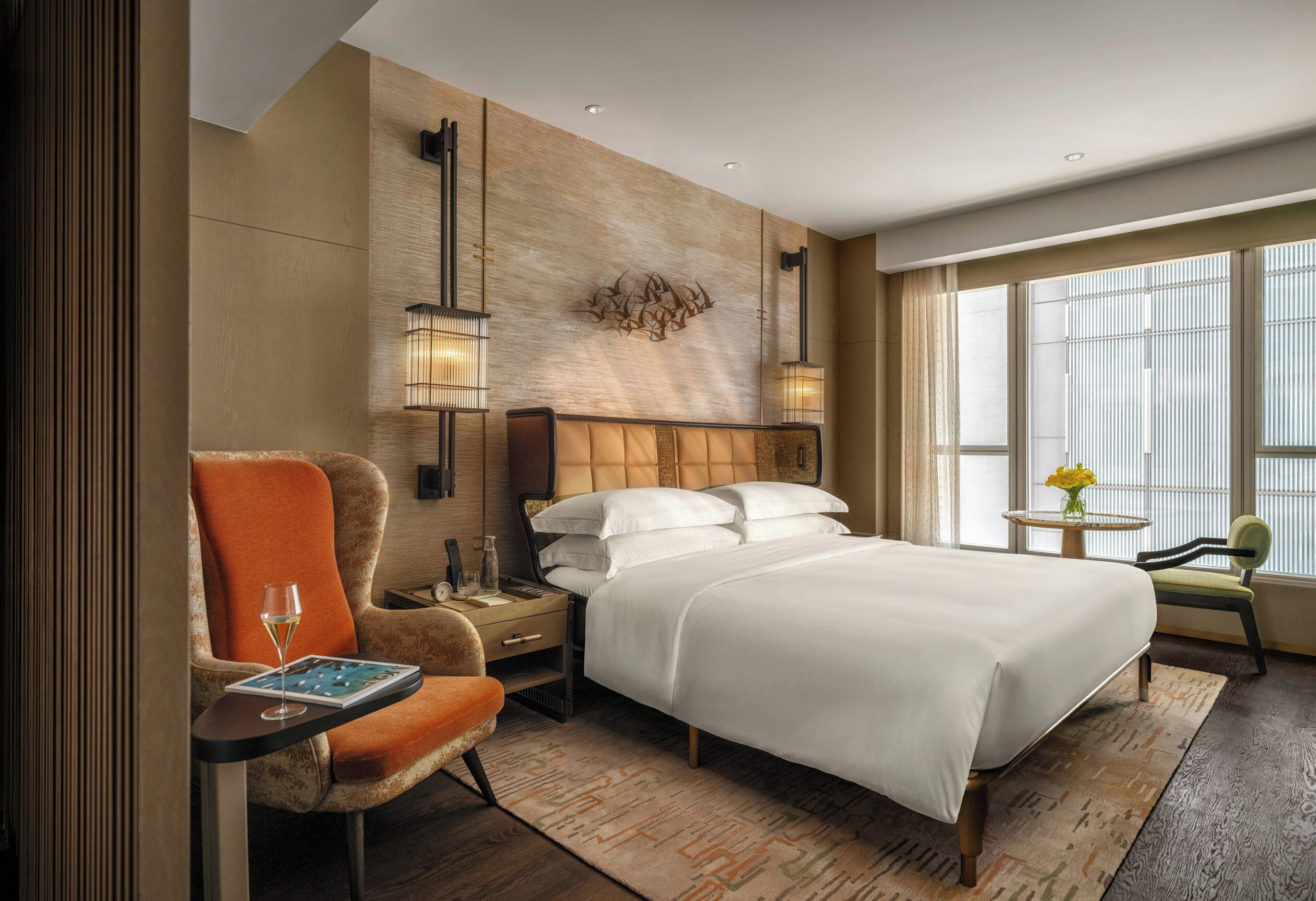 A hotel room with a large bed, a chair, a small table with a glass of wine, a nightstand, and a wall-mounted lamp.