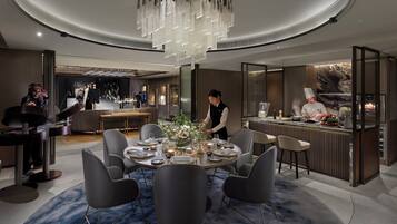 Ein moderner Essbereich mit einem runden Tisch, grauen Stühlen und einer Deckenleuchte. Ein Koch bereitet Essen an einer Theke zu.