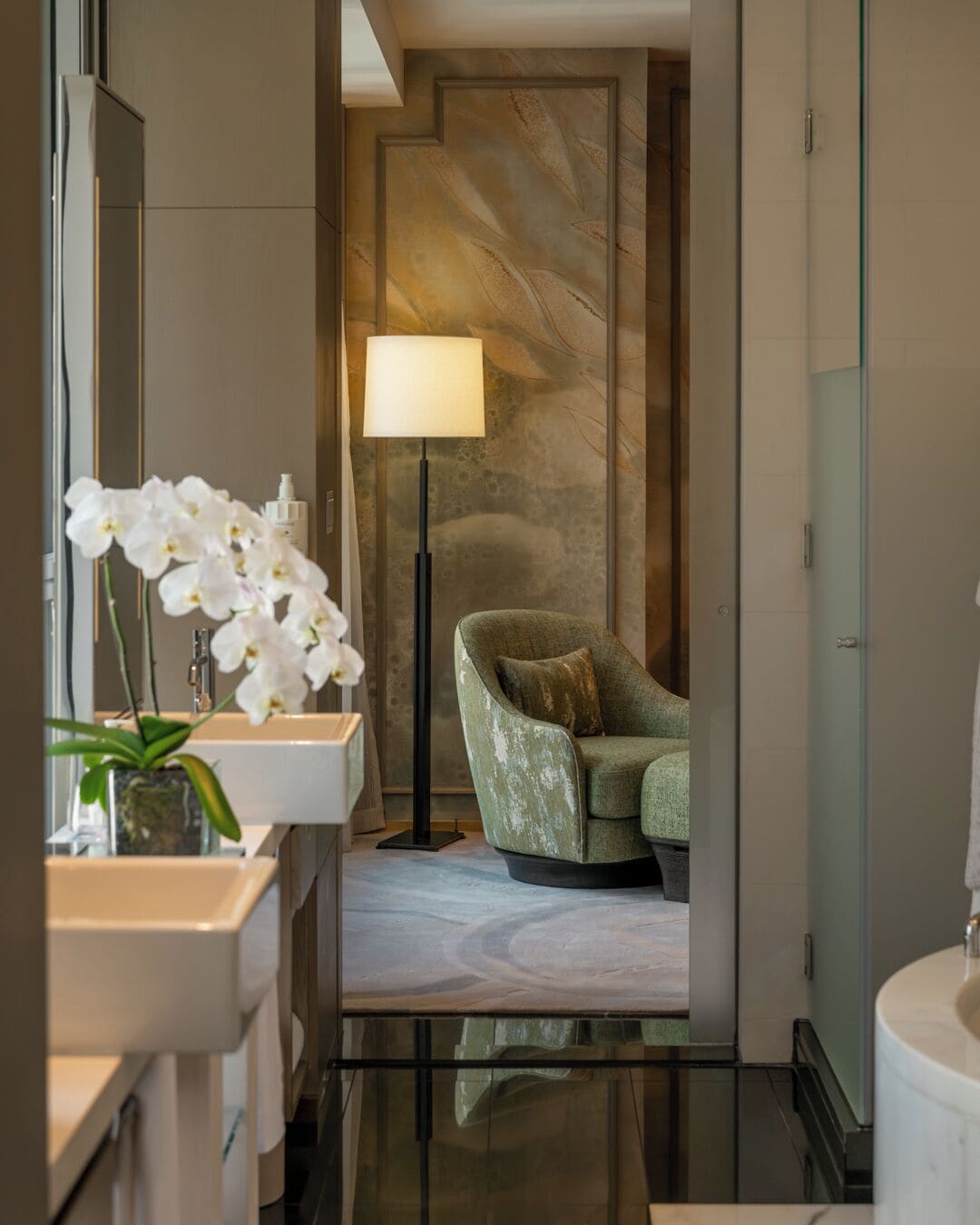 A modern bathroom with a marble floor, a green armchair, a floor lamp, and white orchids.