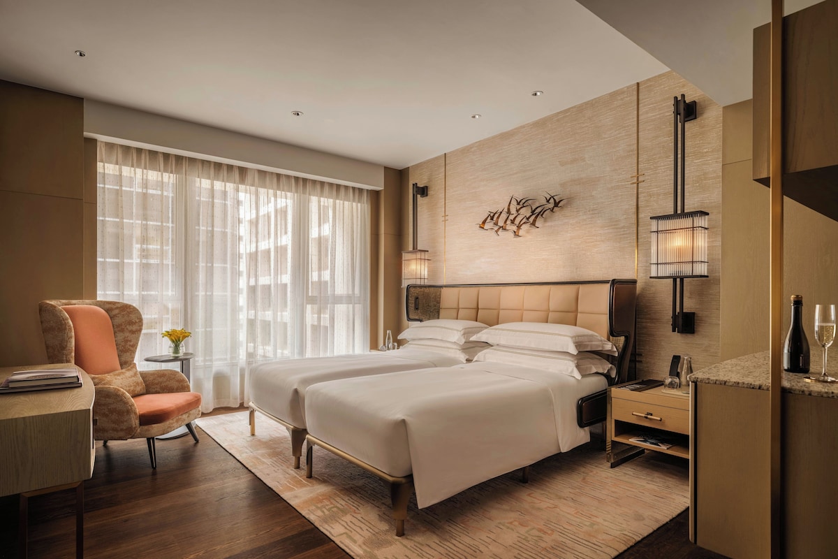 Une chambre d’hôtel moderne avec un lit double, un bureau avec une chaise, une petite table avec un vase et une grande fenêtre avec des rideaux légers.