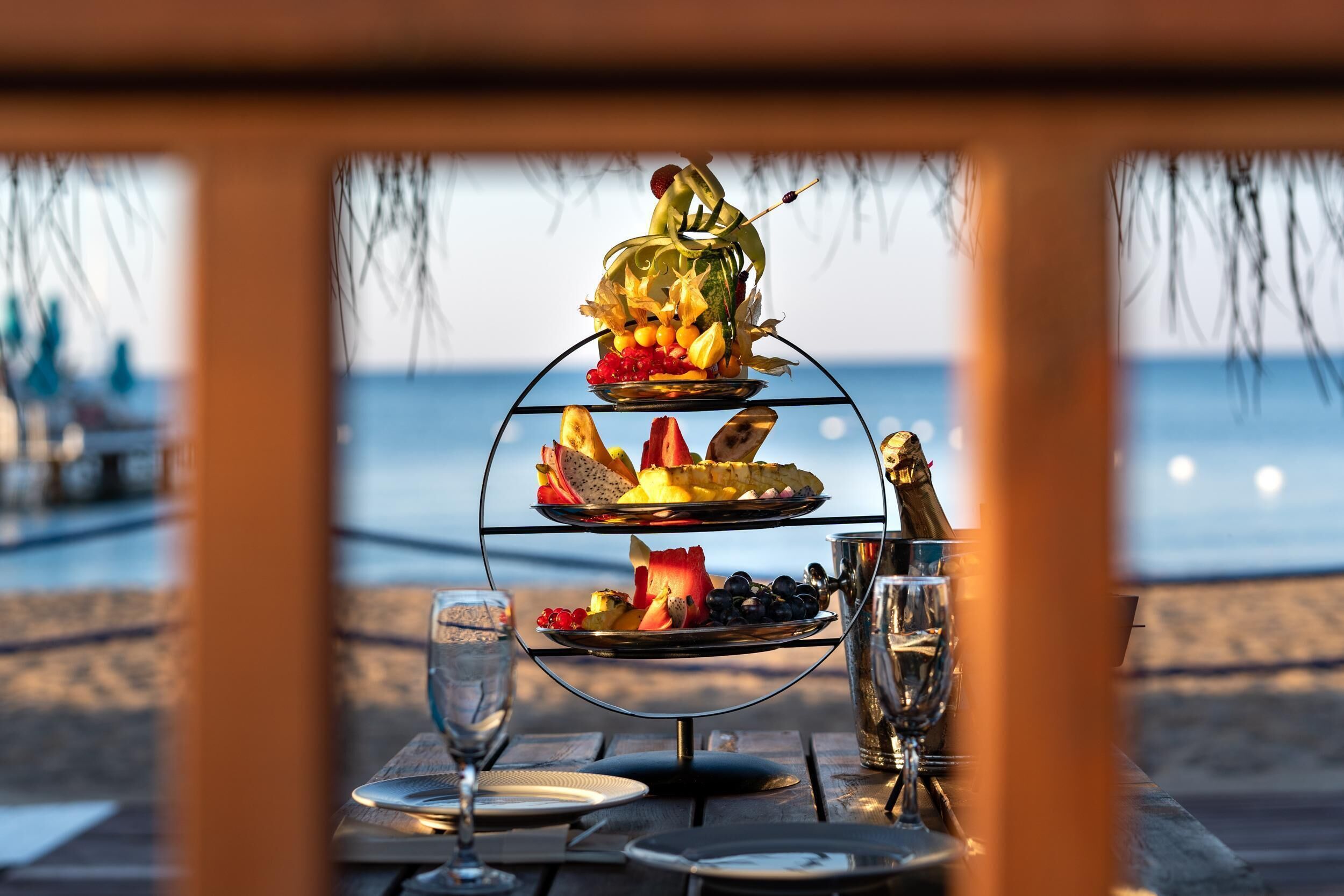 A three-tiered fruit and cheese board with a bottle of champagne, set on a table with a view of the beach and ocean.