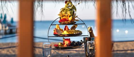 A three-tiered fruit and cheese board with a bottle of champagne, set on a table with a view of the beach and ocean.