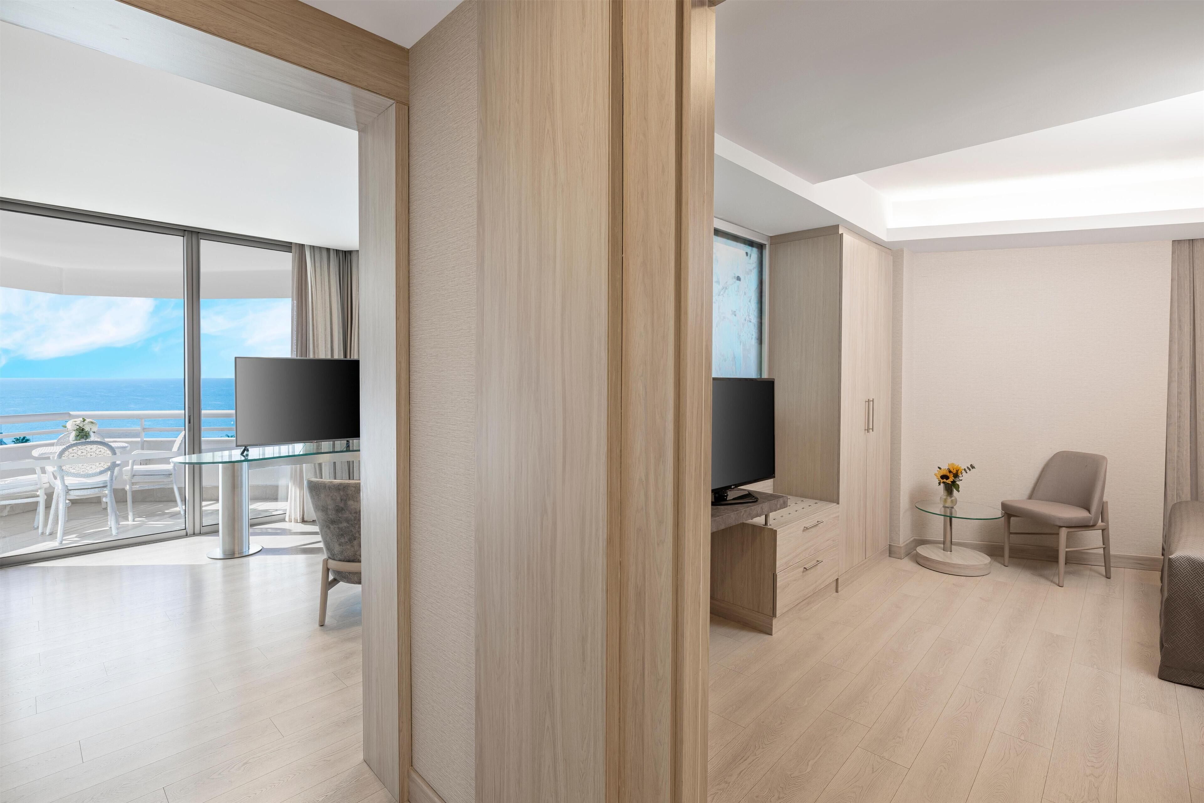 Una habitación de hotel moderna con balcón, un televisor de pantalla plana, una pequeña mesa redonda con un florero y una silla cómoda.