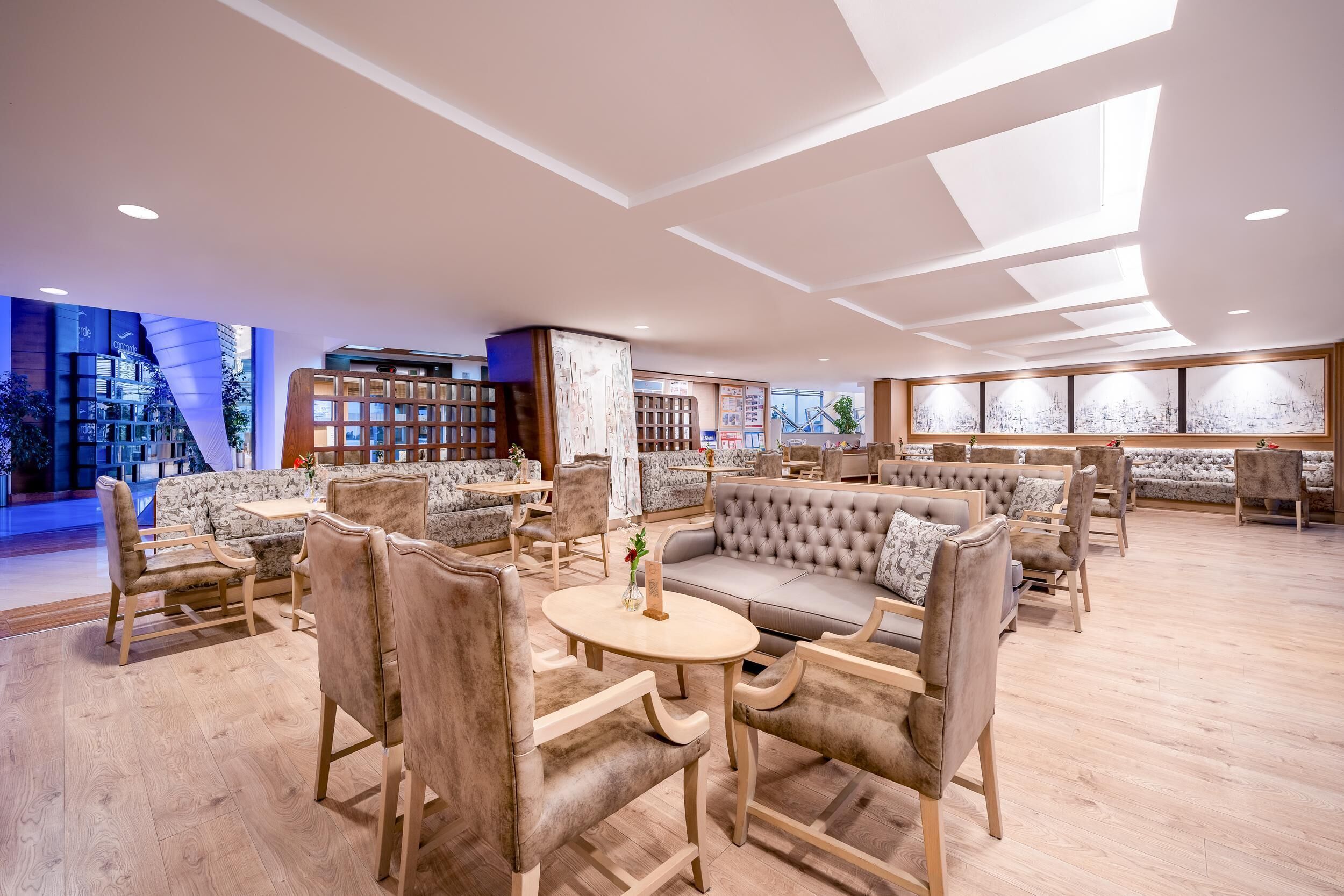 A spacious lounge area with sofas, chairs, and a round table, featuring a modern ceiling design and large windows.