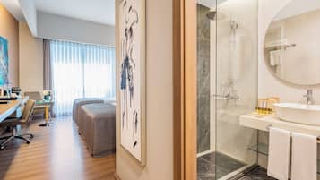 A modern hotel room with a wooden floor, a glass shower enclosure, a white sink, and a large mirror. There is a bed with a grey headboard and a small table with a vase of flowers.