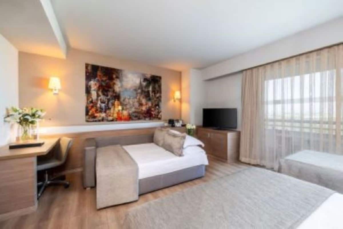 A modern hotel room with a bed, a desk, a chair, a TV, and a large painting on the wall.