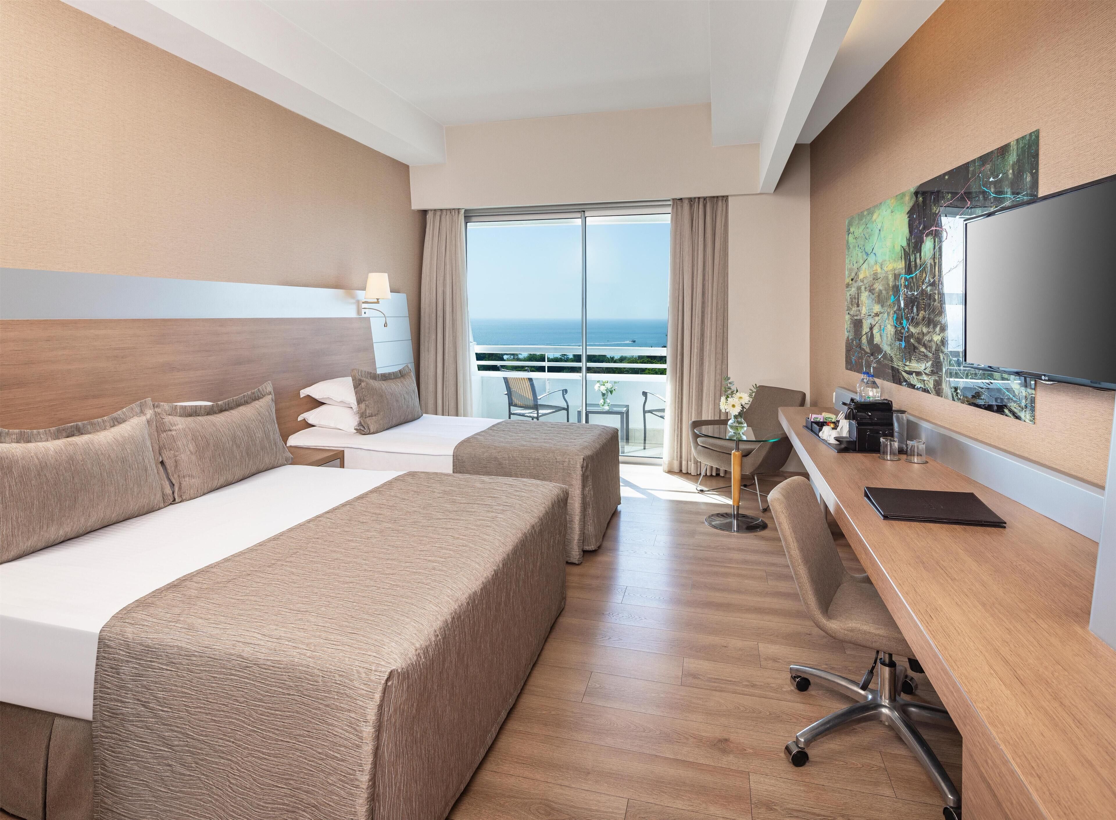 A hotel room with two beds, a desk, a chair, a TV, and a balcony with a view of the sea.