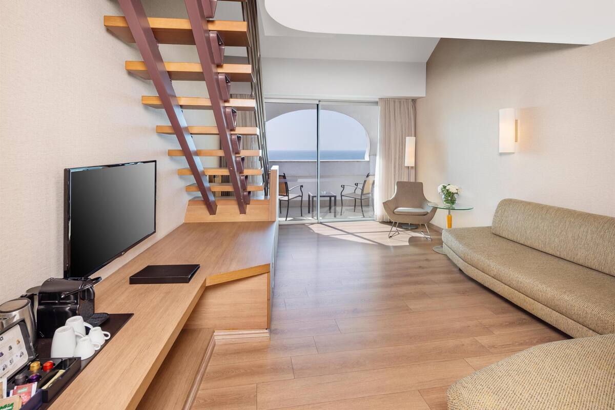 A modern hotel room with a wooden staircase, a flat-screen TV, a sofa, and a balcony with a view of the sea.