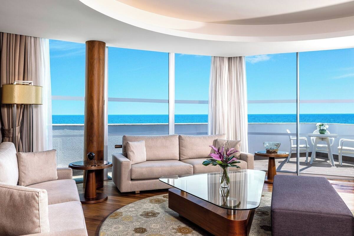A modern living room with a large glass table, beige sofas, and a view of the ocean.