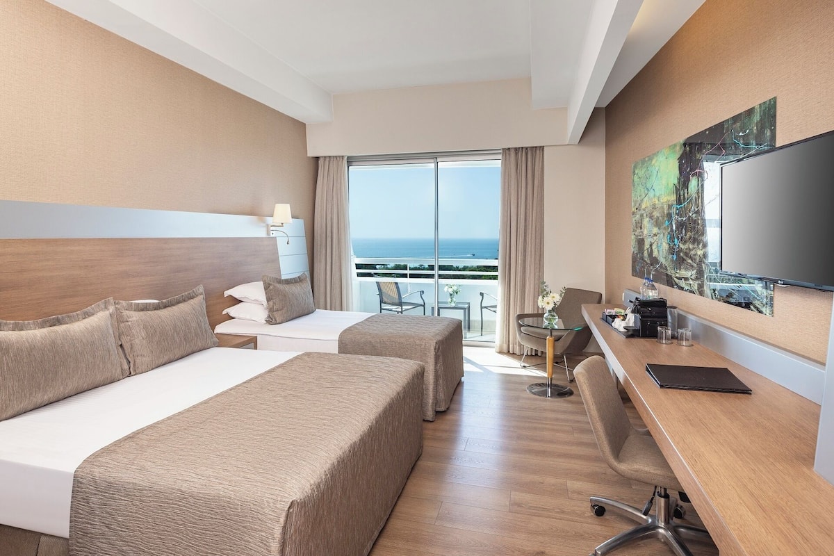A hotel room with two beds, a desk, a chair, a TV, and a balcony with a view of the ocean.
