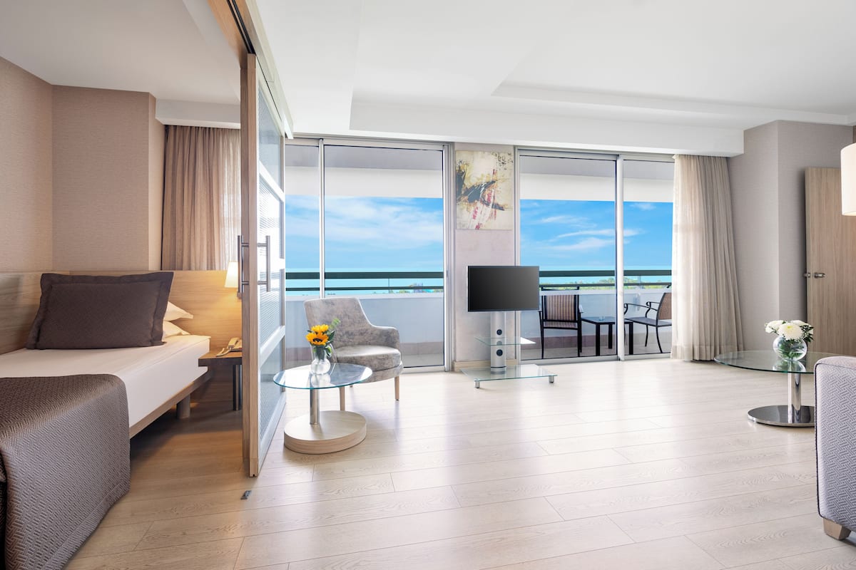 A modern hotel room with a balcony, a bed, a sofa, a TV, and a dining area with a view of the ocean.