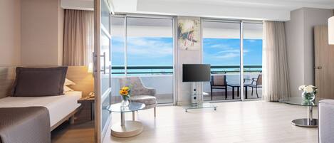 A modern hotel room with a balcony, a bed, a sofa, a TV, and a dining area with a view of the ocean.