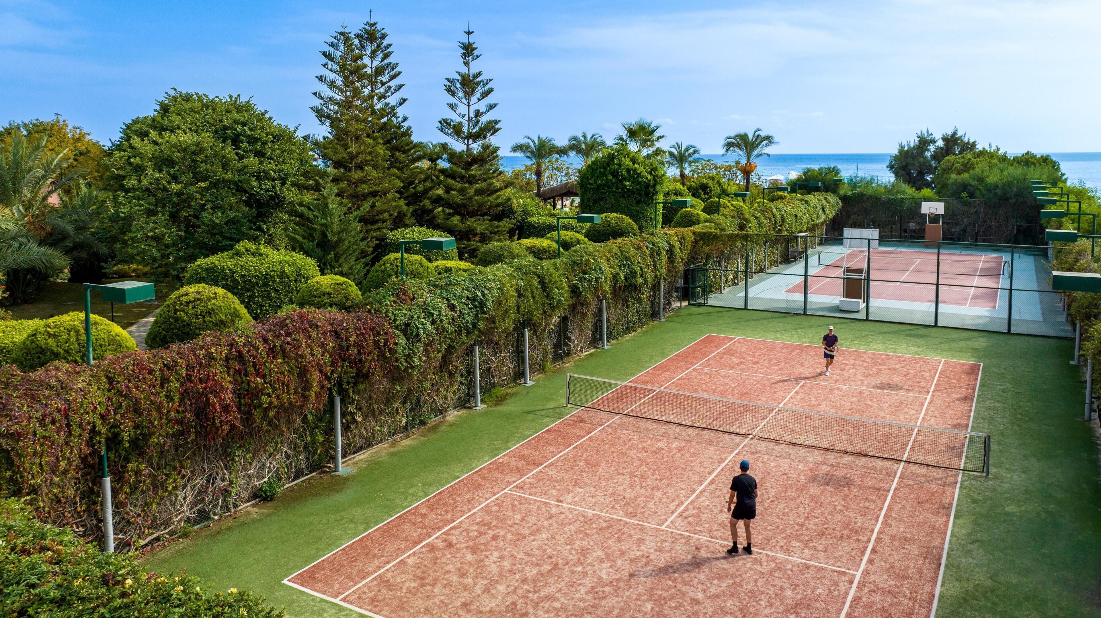 A tennis court with a net, surrounded by lush greenery and a view of the sea.