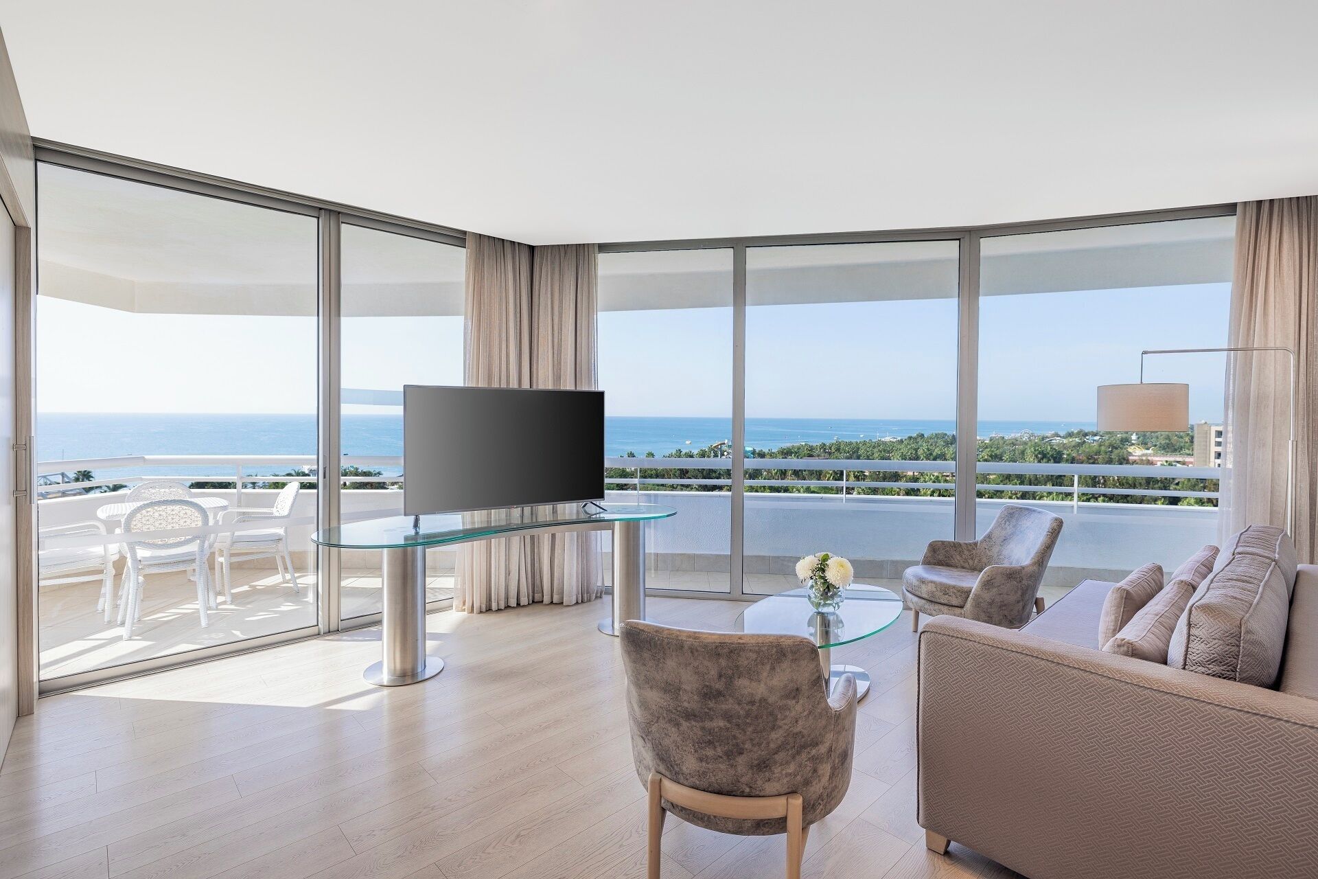 A modern hotel room with a large balcony, a flat-screen TV, a glass coffee table, and a sofa.