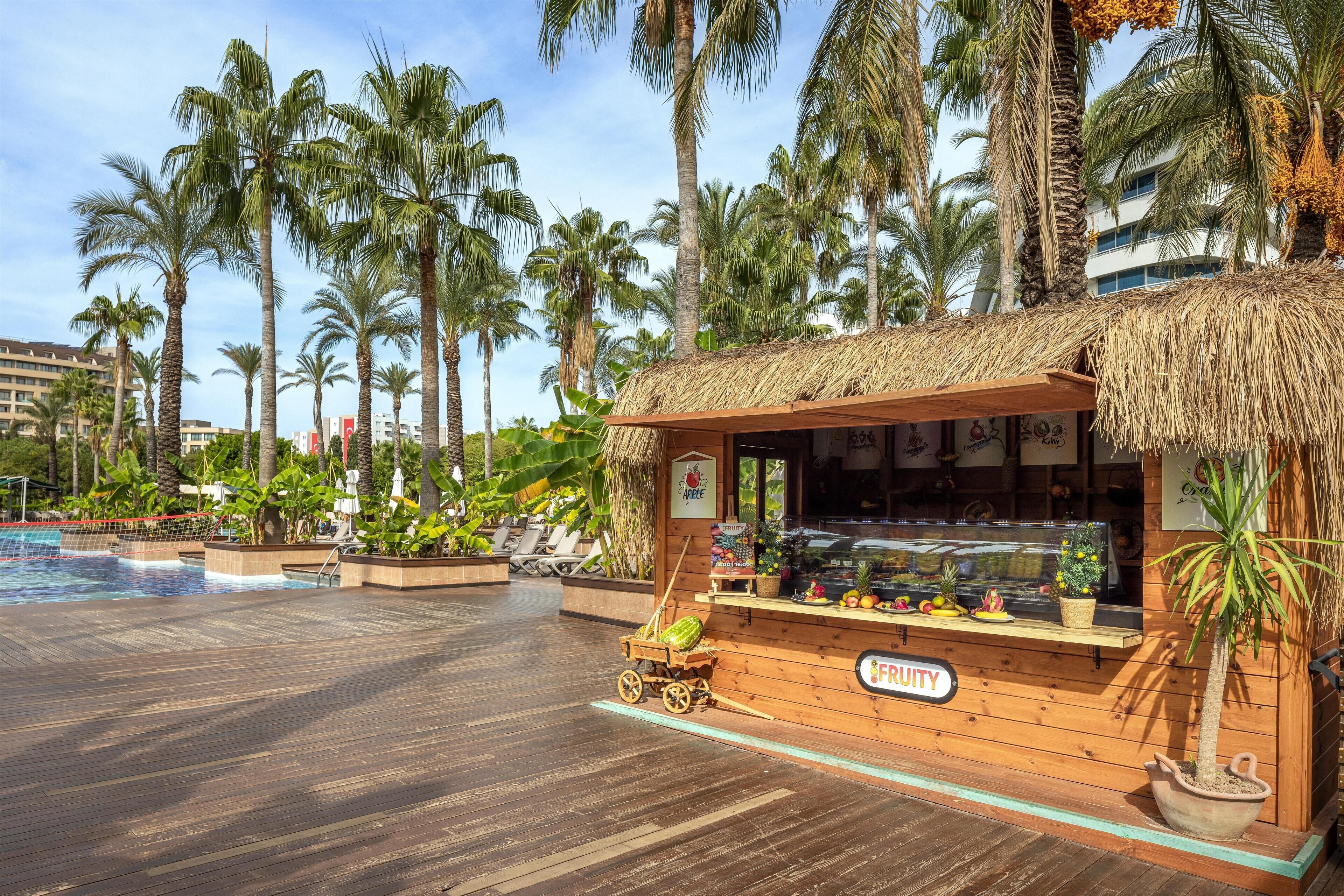A wooden kiosk with a thatched roof, a sign reading "FRUITY," and a display of fruits and vegetables.