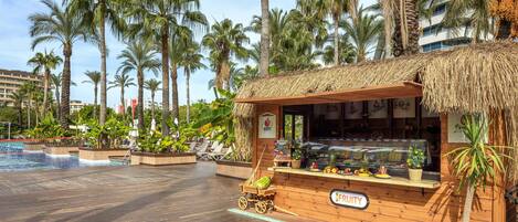 A wooden kiosk with a thatched roof, a sign reading "FRUITY," and a display of fruits and vegetables.