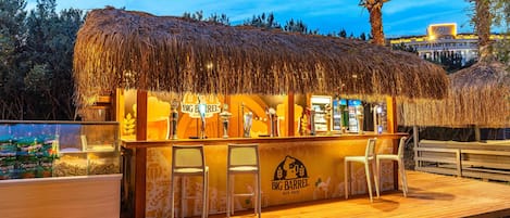 Un'area bar all'aperto con tetto in paglia, sgabelli da bar e un cartello che recita "Big Barrel Beer House".