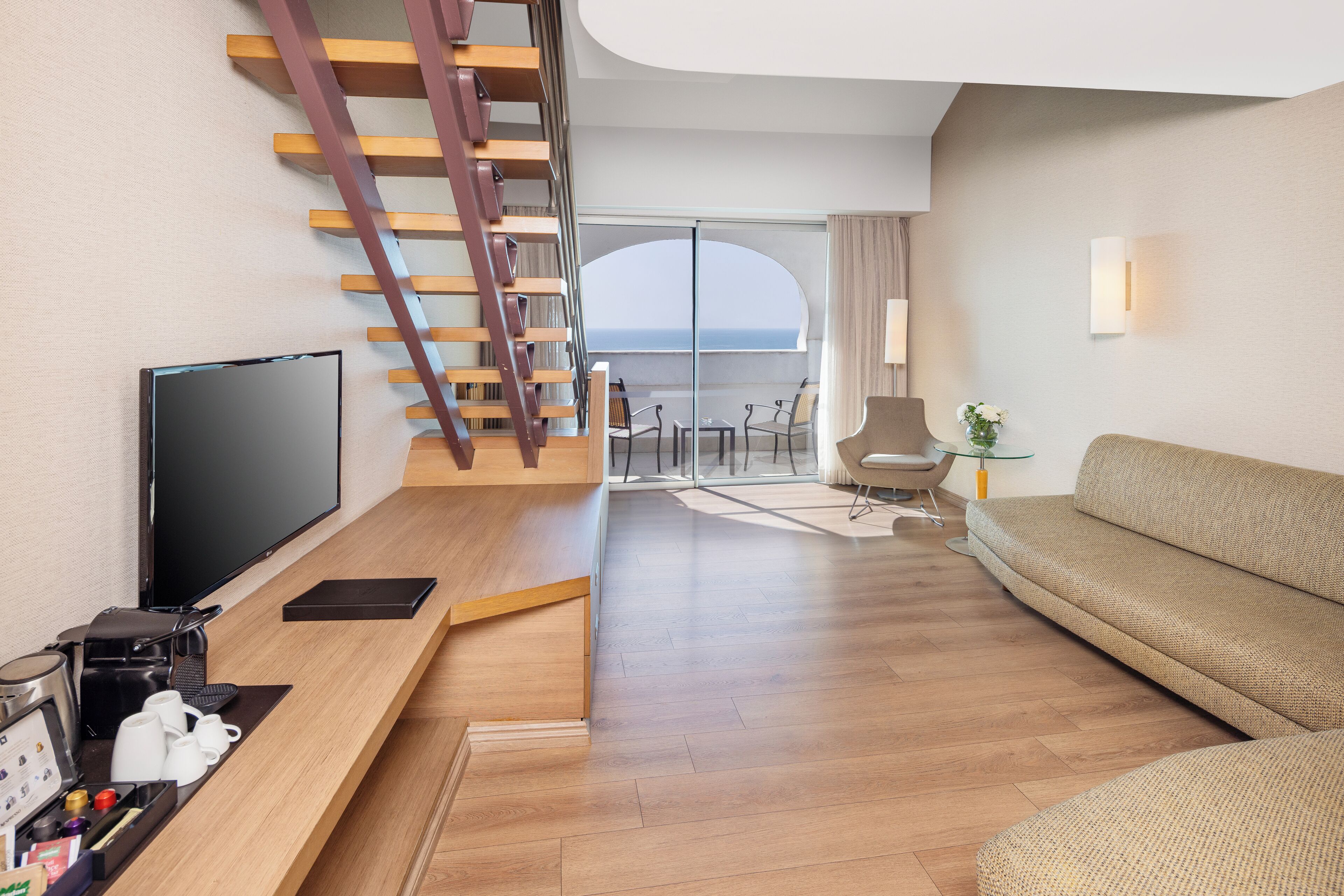 Habitación de hotel moderna con escalera de madera, televisor de pantalla plana, sofá y balcón con vista al mar.
