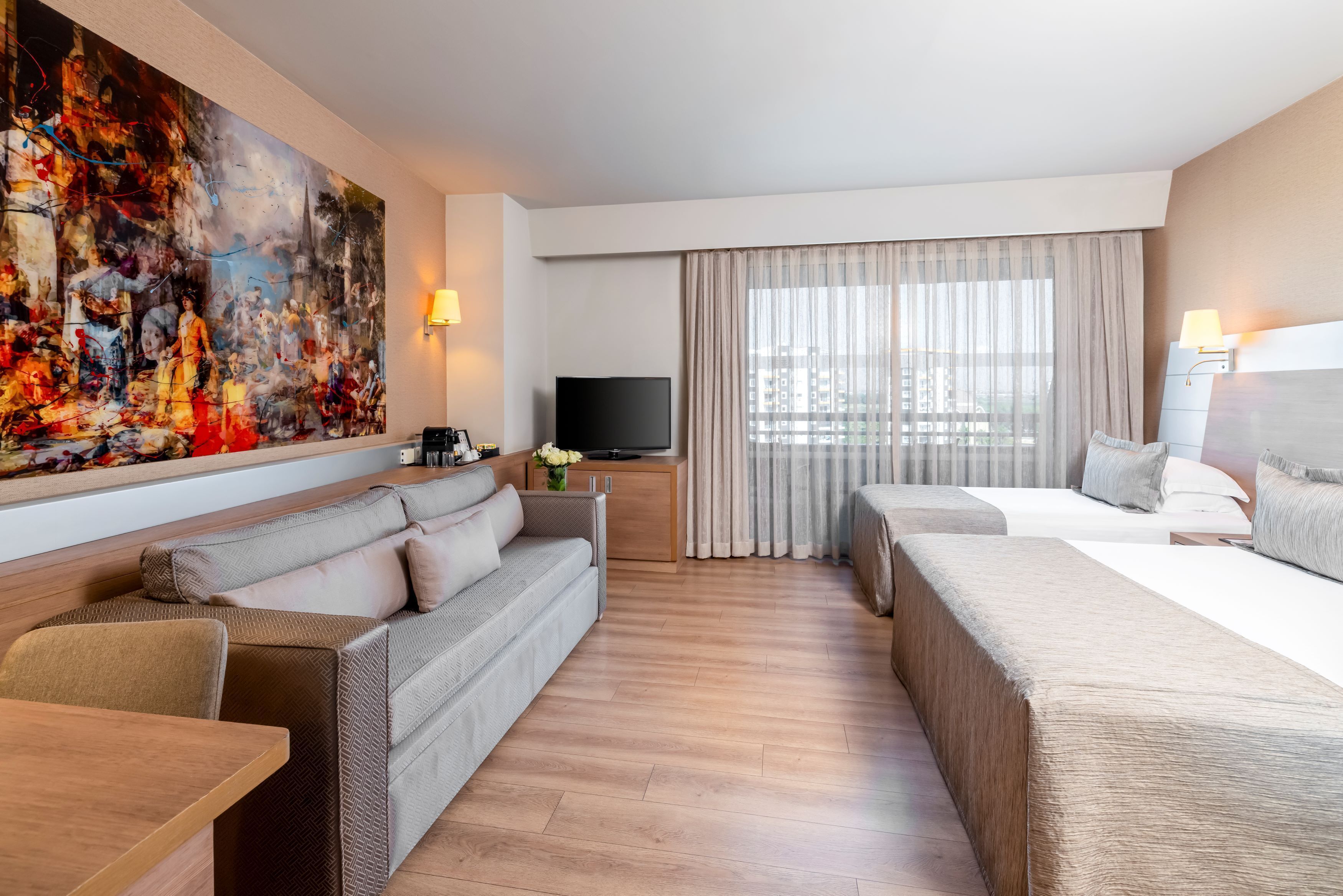 A modern hotel room with a large abstract painting, a sofa, a bed, a TV, and a view of the city.