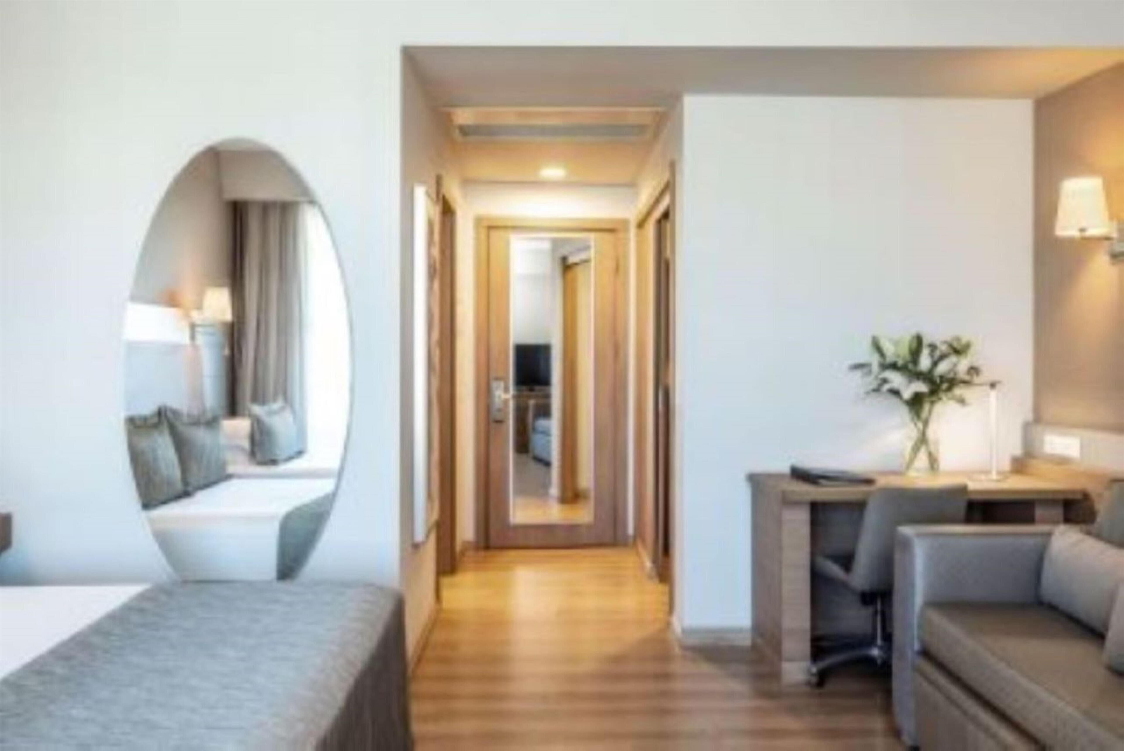A hotel room with a bed, a desk, and a chair. There is a mirror on the wall and a plant on the desk.