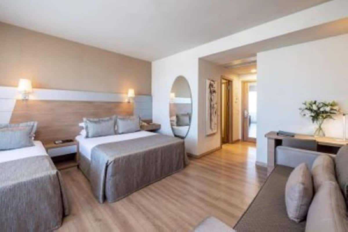 A modern hotel room with two single beds, a sofa, a desk, and a mirror.