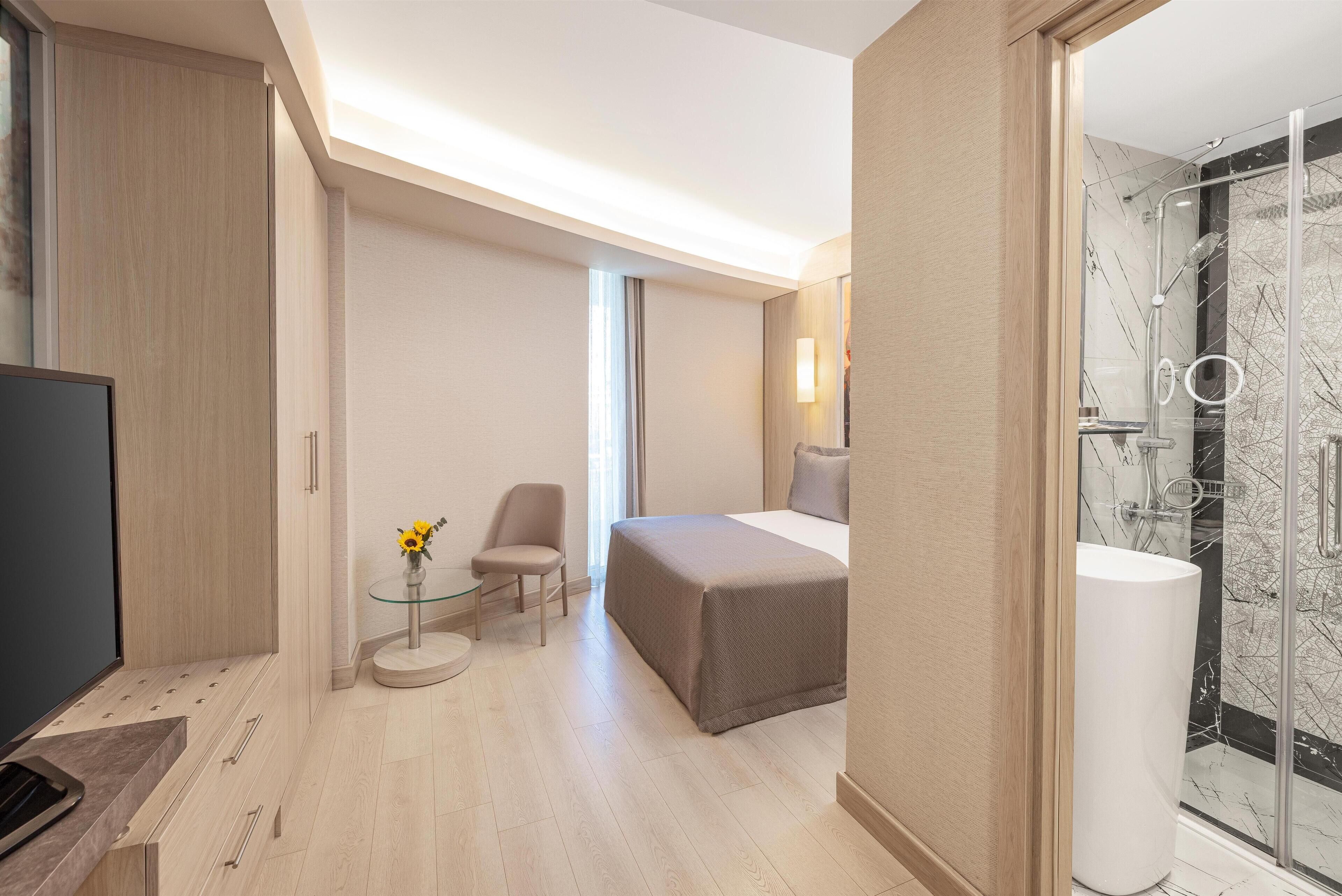 A modern hotel room with a bed, a TV, a small table with a vase, a chair, and a shower area.
