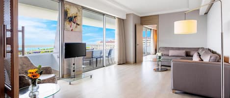 A modern hotel room with a large balcony, a flat-screen TV, a sofa, a coffee table, and a lamp.