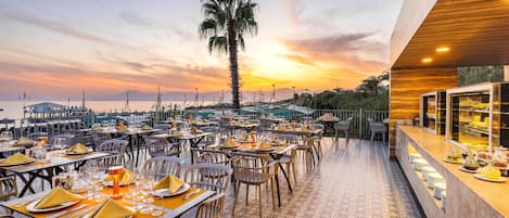 An outdoor dining area with tables set for a meal, overlooking a body of water and a sunset.