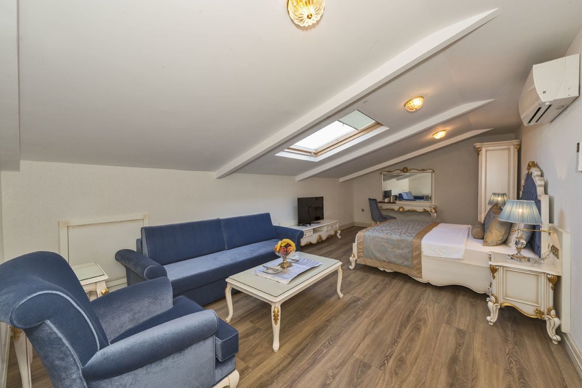 Uma sala de estar ampla com um sofá azul, uma mesa de centro e uma televisão. Há um quarto com uma cama e um espelho. O ambiente possui piso de madeira e um claraboia.