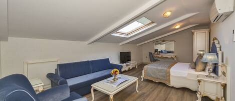 Uma sala de estar ampla com um sofá azul, uma mesa de centro e uma televisão. Há um quarto com uma cama e um espelho. O ambiente possui piso de madeira e um claraboia.
