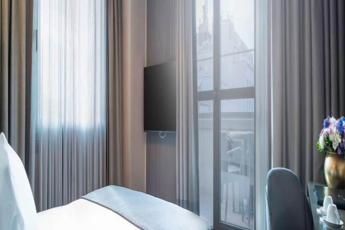 A hotel room with a bed, a flat-screen TV mounted on the wall, a desk with a vase of flowers, and a view of buildings through the window.