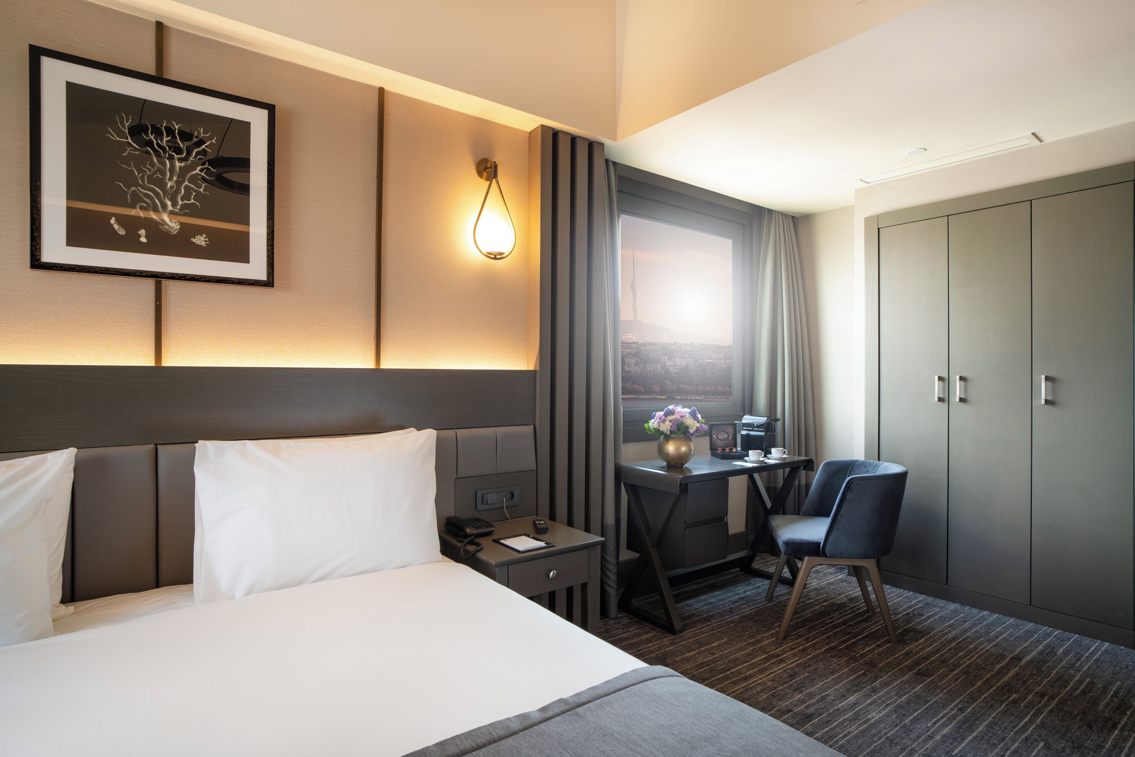 A hotel room with a bed, a desk with a chair, a wardrobe, a framed picture on the wall, and a window with curtains.