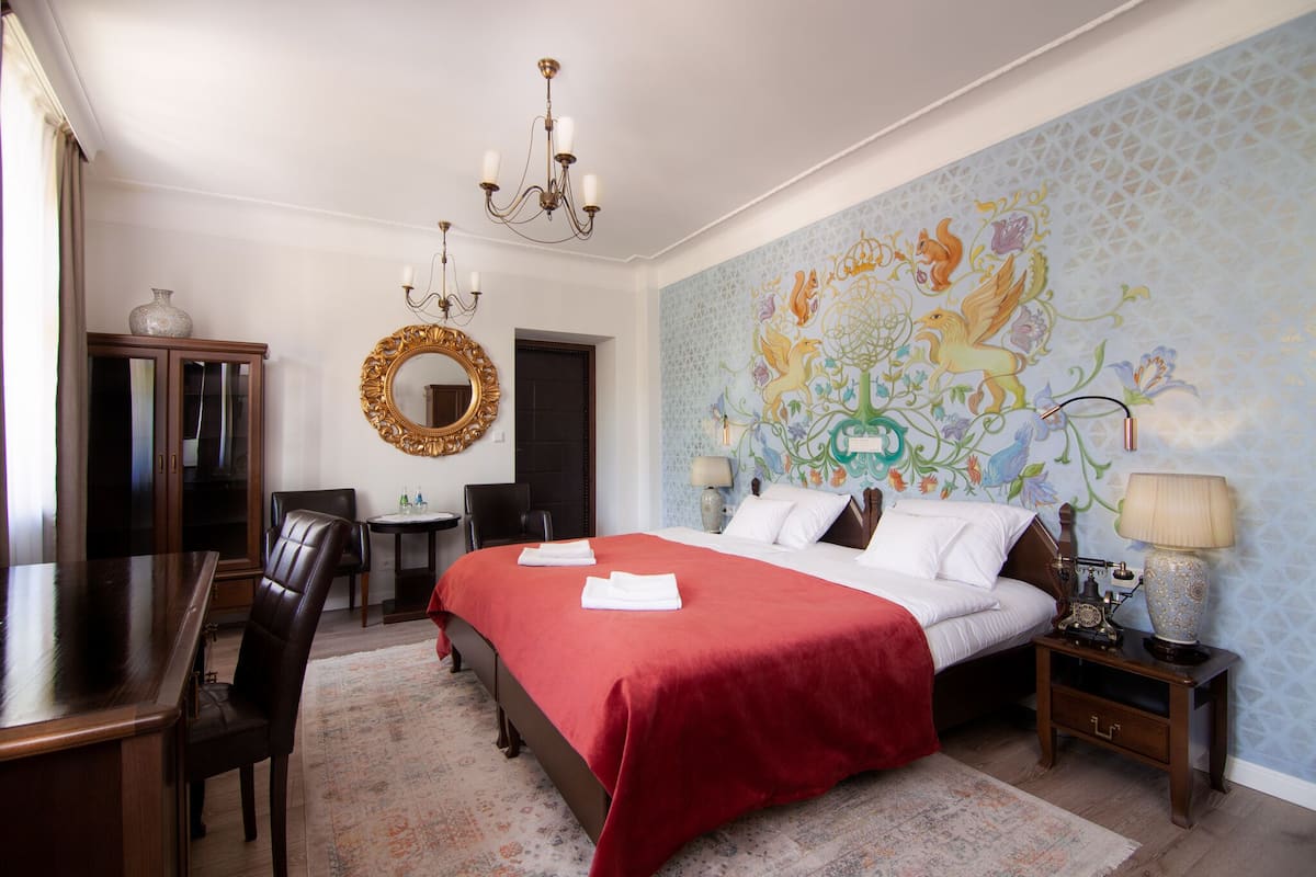Habitación de hotel con una cama grande, un escritorio de madera, una araña, un espejo y un mural decorativo en la pared.