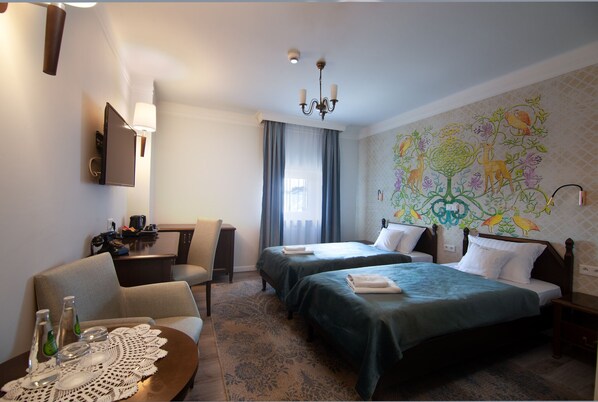Quarto de hotel com duas camas, uma escrivaninha, uma cadeira, uma mesinha com um bordado e uma parede com um mural floral.