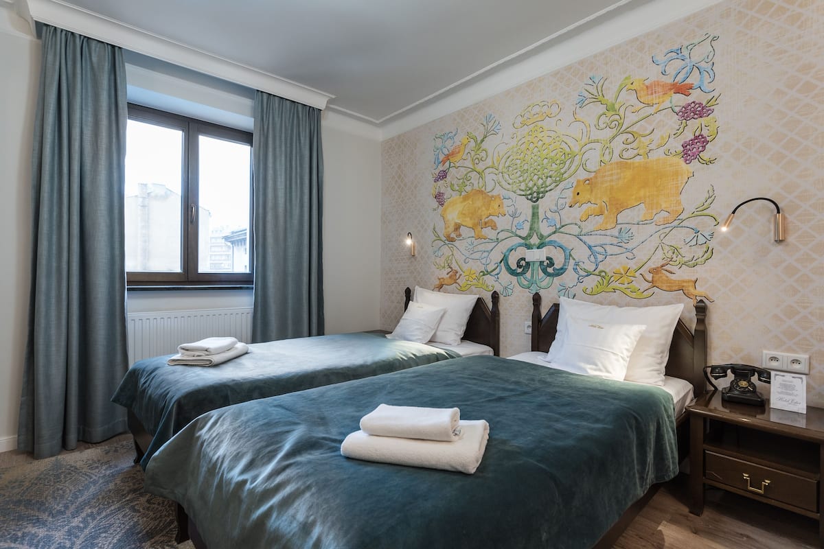 Habitación de hotel con dos camas, una ventana con cortinas, un mural en la pared y una mesita de noche con teléfono.