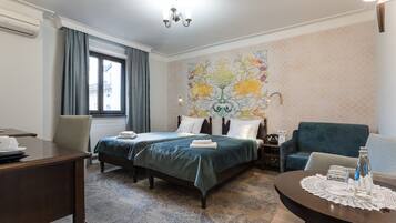 Ett hotellrum med två sängar, ett matbord, en soffa, en dekorativ väggmålning och en kristallkrona.
