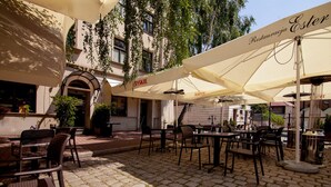 An outdoor café with tables and chairs under umbrellas, a building entrance, and greenery.